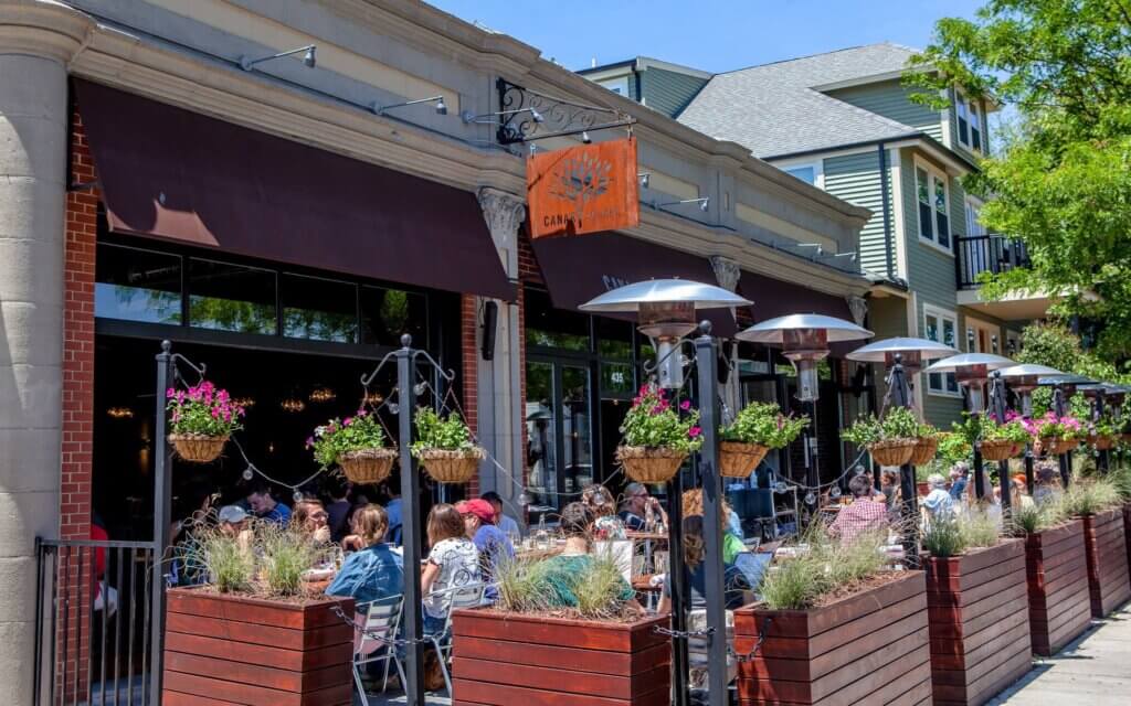 people eating in an outdoor seating area of a restaurant with wood planters and outdoor heating towers around the edges