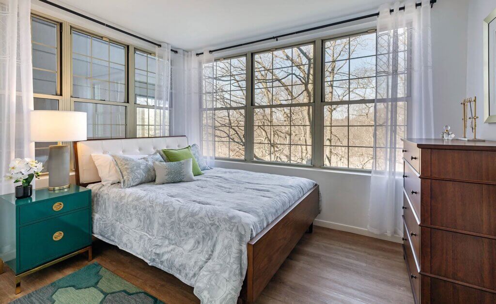 bedroom with a made bed, teal nightstand with lamp and flowers, dark wooden dresser and walls lined with 6 continuous windows